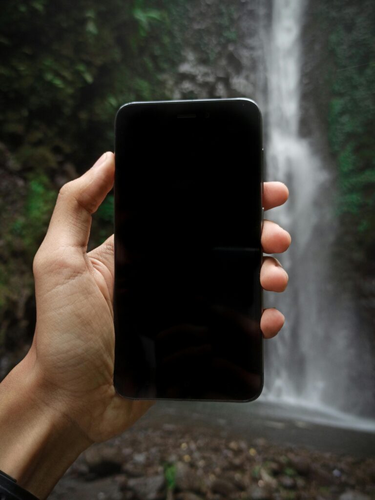 A hand holding a smartphone in front of a blurred waterfall, capturing nature's beauty.